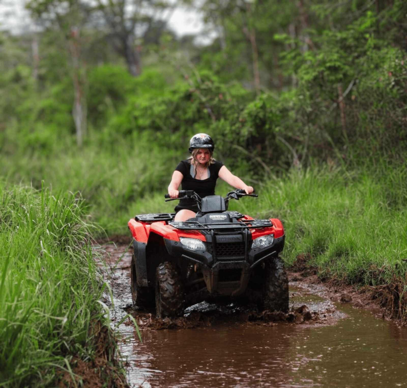 A young rider in a helmet steering a red ATV through a muddy puddle between banks of tall green grass and forest