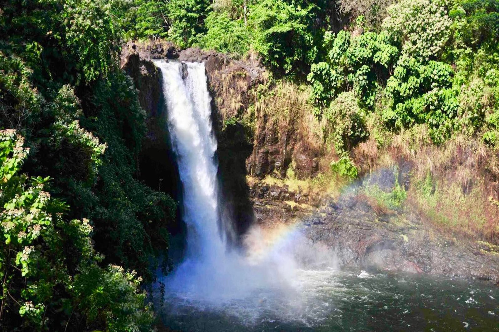 A waterfall plunging over a dark lava ledge into a churning pool, with a faint rainbow in the mist and green forest all around