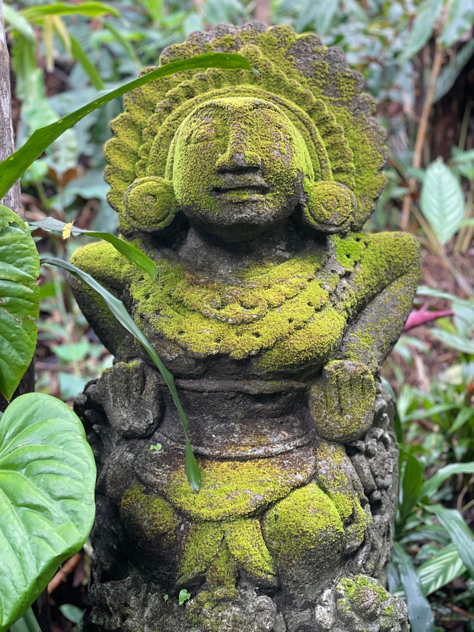 A stone statue covered in bright green moss, half hidden among leaves at the side of the trail