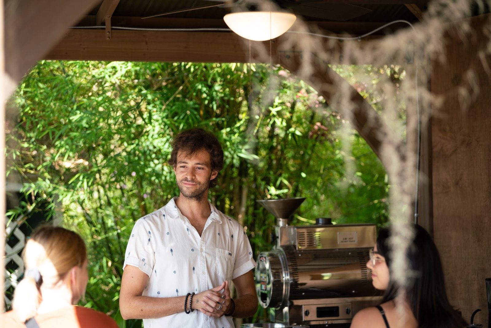A brew master in a white shirt talking with guests under a covered lanai, with a coffee roaster and green bamboo behind him