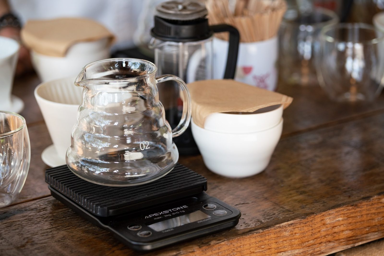 A glass carafe on a digital scale beside paper filters, white cupping bowls and a French press on a wooden bar