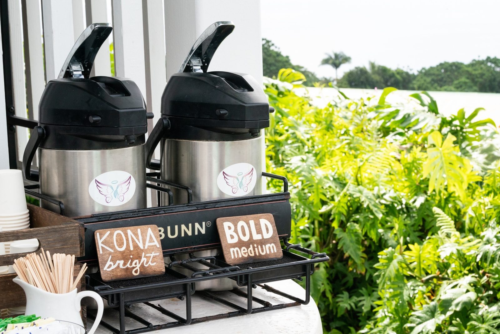 Two coffee dispensers labeled Kona bright and bold medium on a sampling stand beside tropical plants