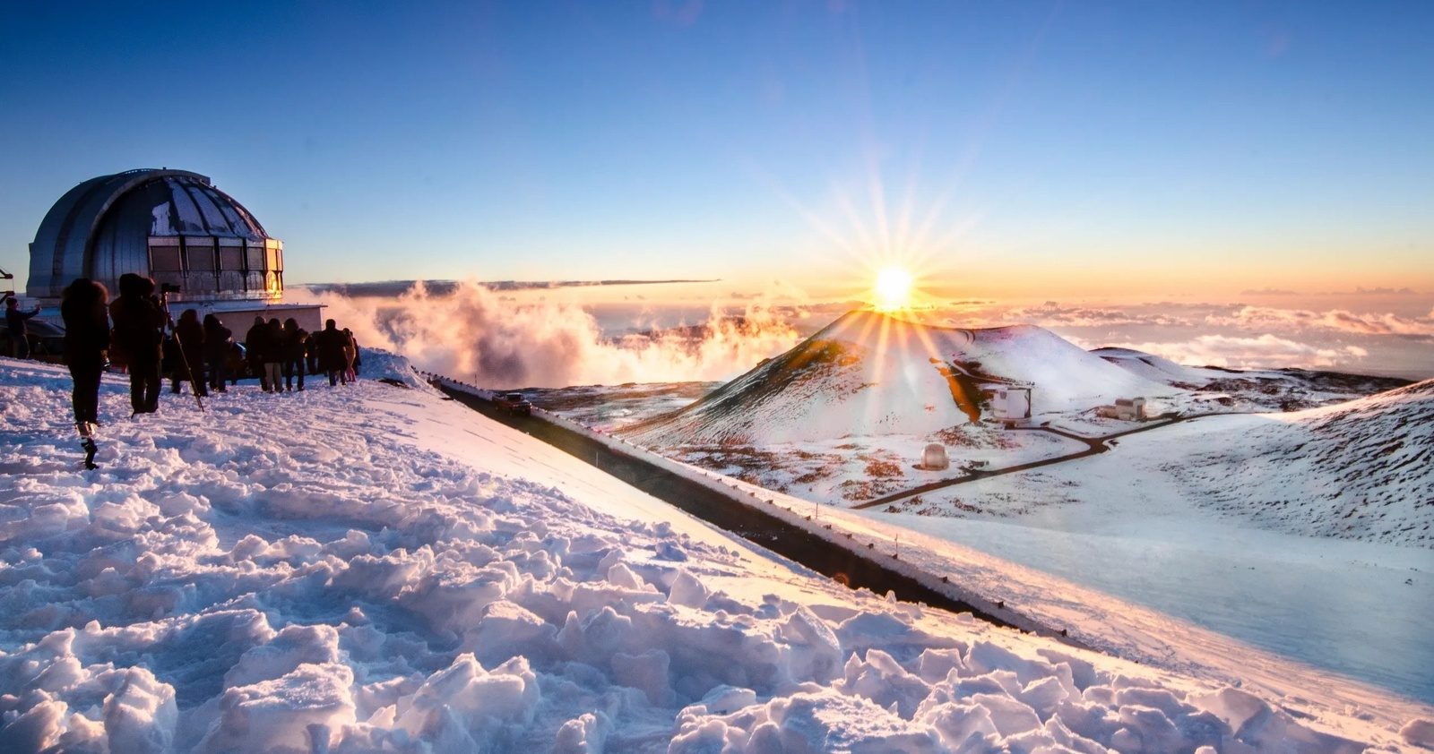 Visitors in heavy jackets standing in snow beside a silver observatory dome, watching the sun set over a cinder cone and a layer of clouds