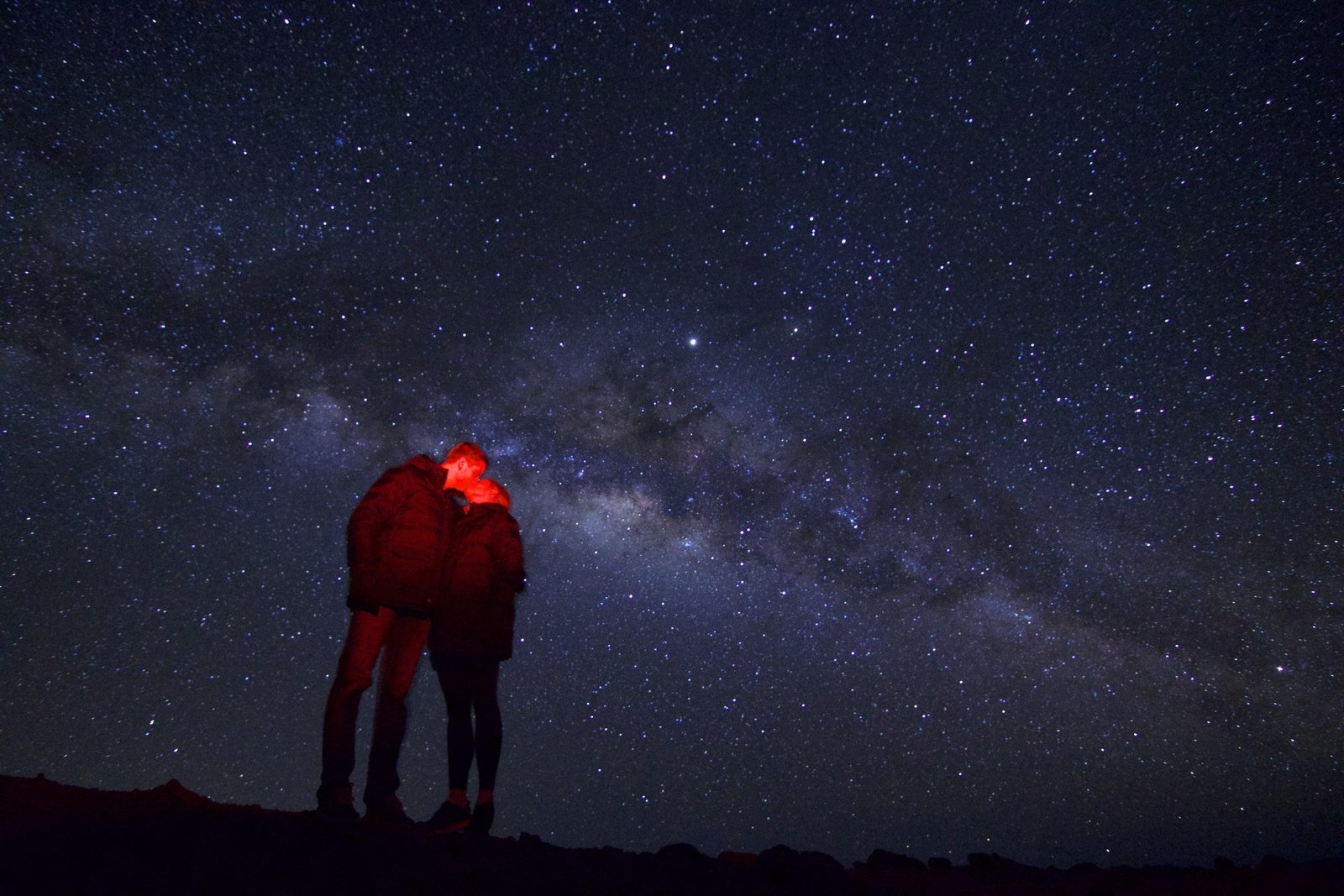 A couple in puffy jackets sharing a kiss under the Milky Way, lit softly in red light
