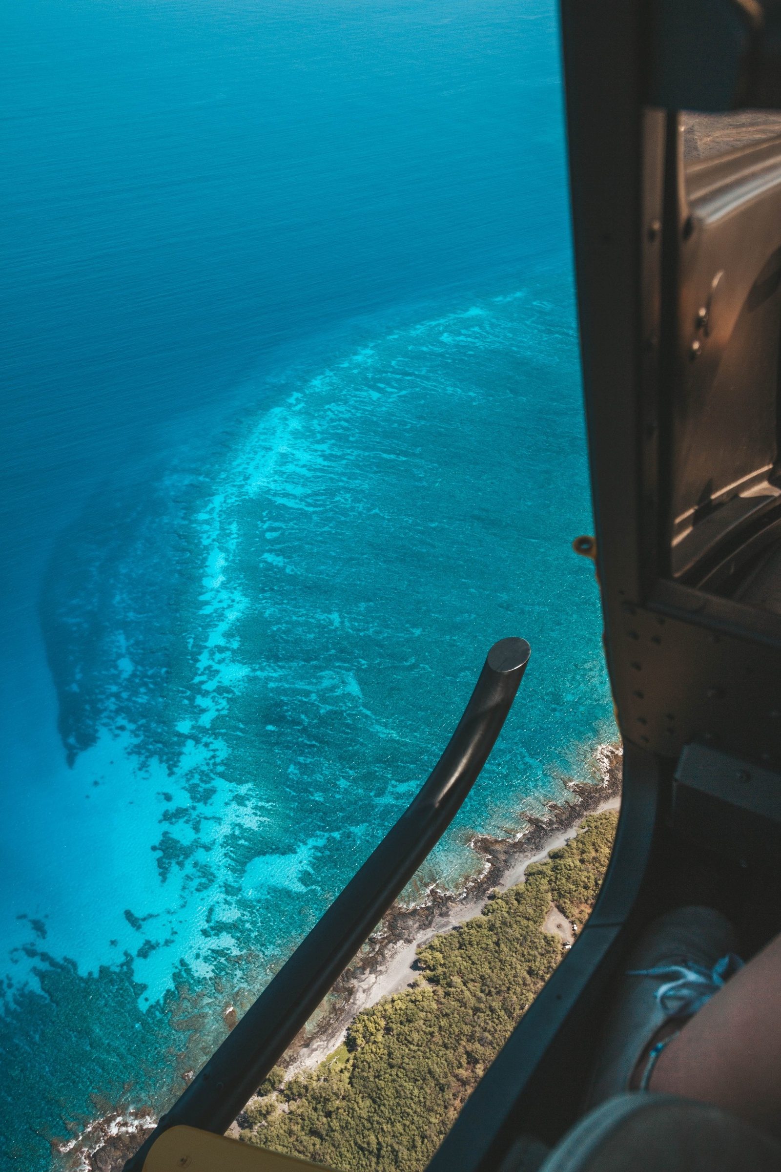 Looking straight down past the landing skid at a fringing reef in bright blue water off a dry, scrubby shoreline