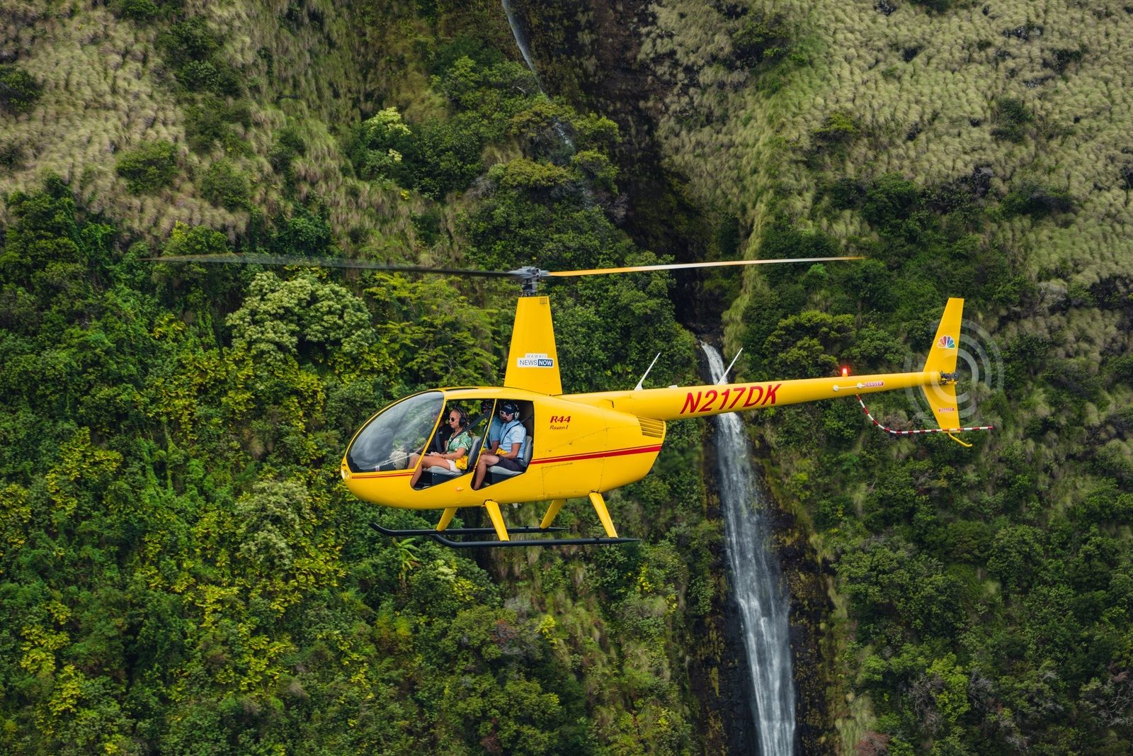 A yellow four-seat helicopter hovering in front of a waterfall in a dense green rainforest valley