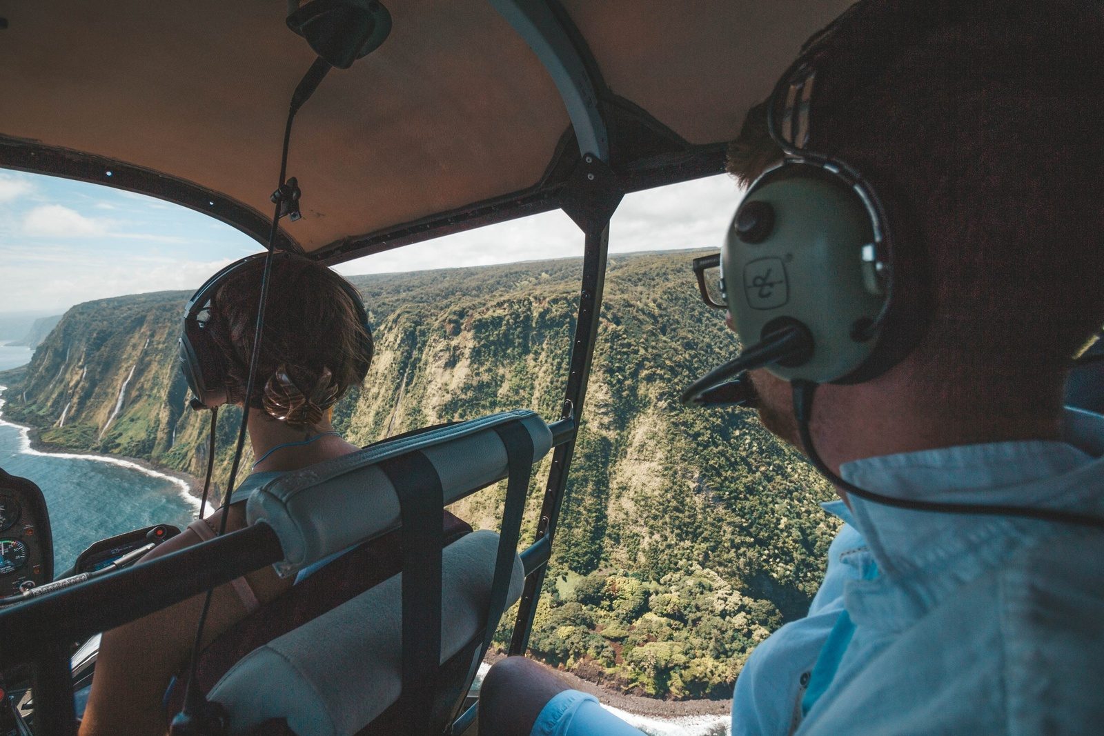Two passengers in headsets looking out the side window of the helicopter at a deep green valley wall and the coastline below