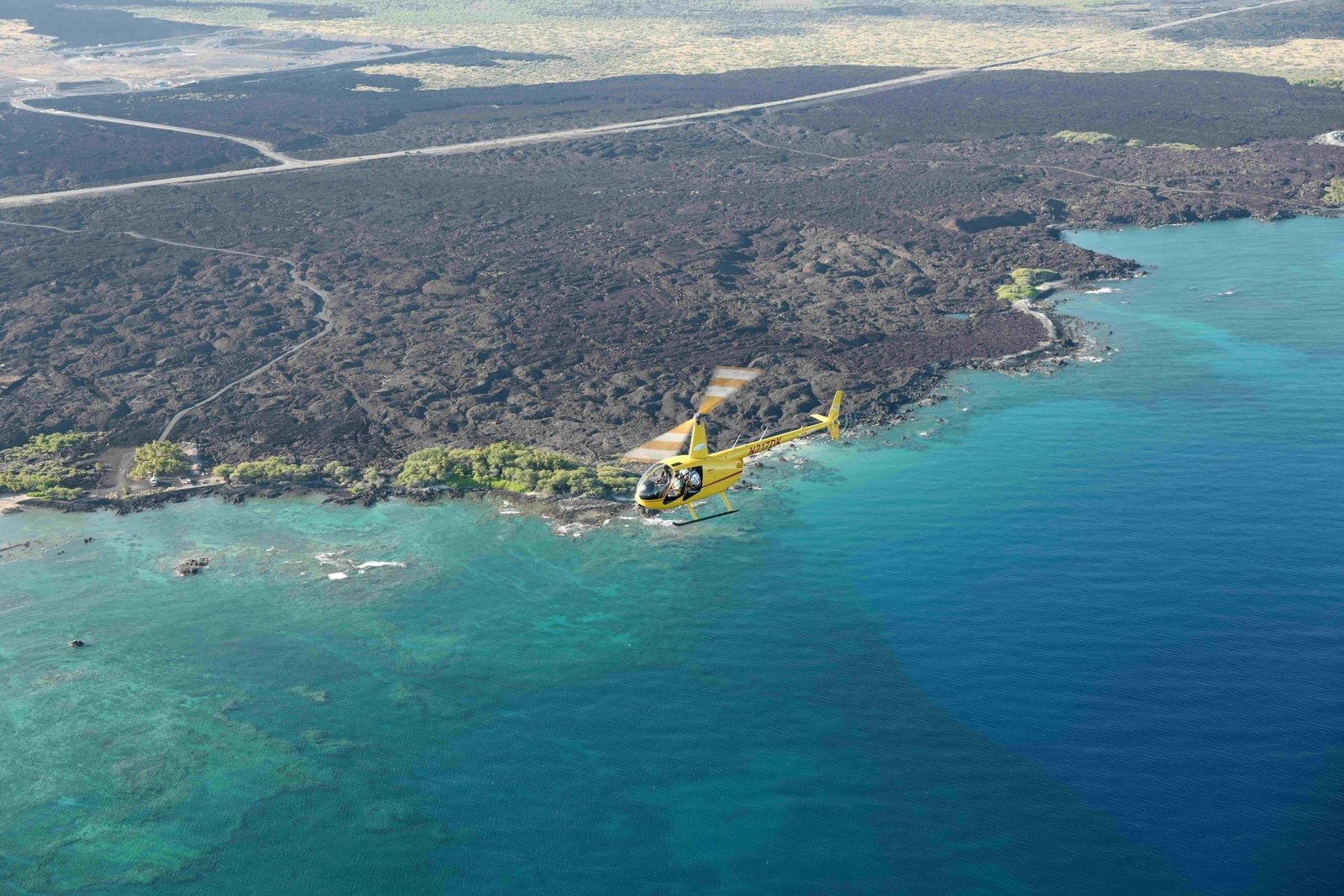 The yellow helicopter flying over turquoise reef water along a black lava coastline on the dry Kona side