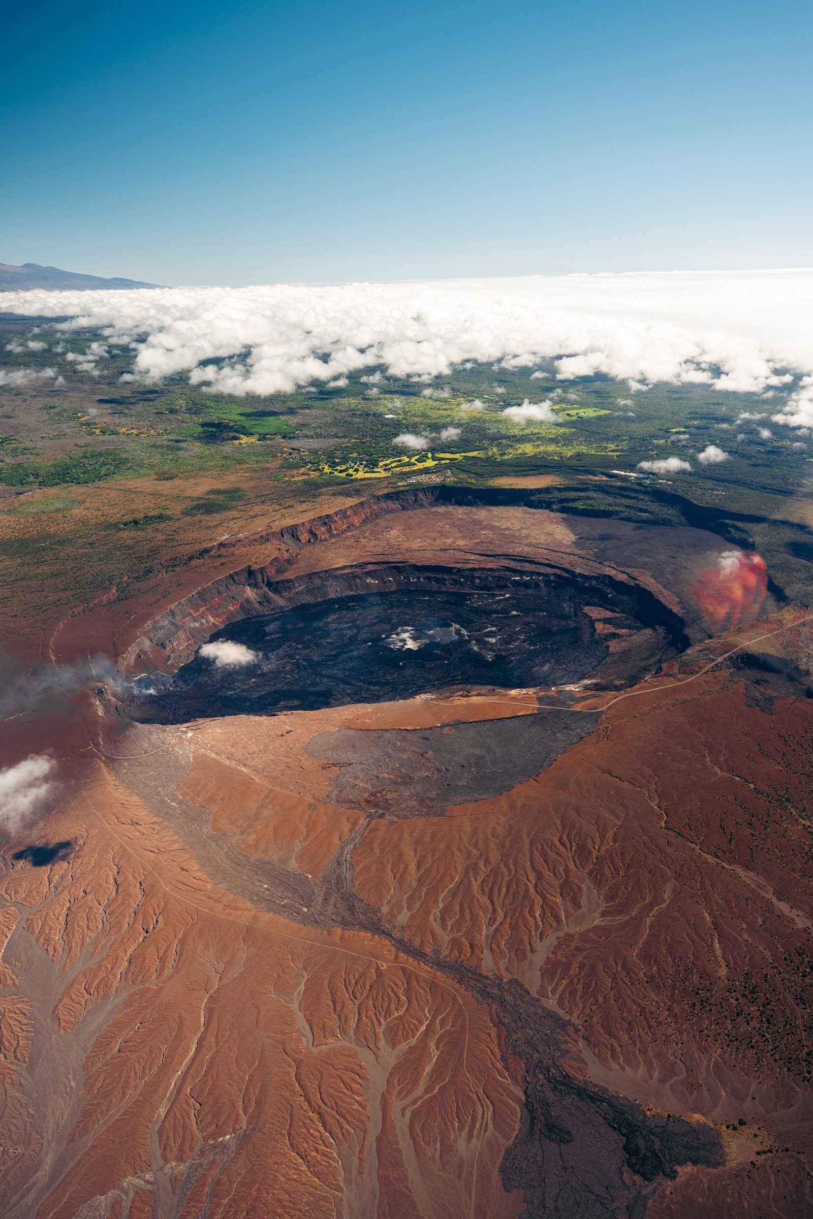 Aerial view of Kīlauea's summit caldera, a vast crater floored with dark lava and rimmed by rust-red ash, with clouds and Maunakea on the horizon