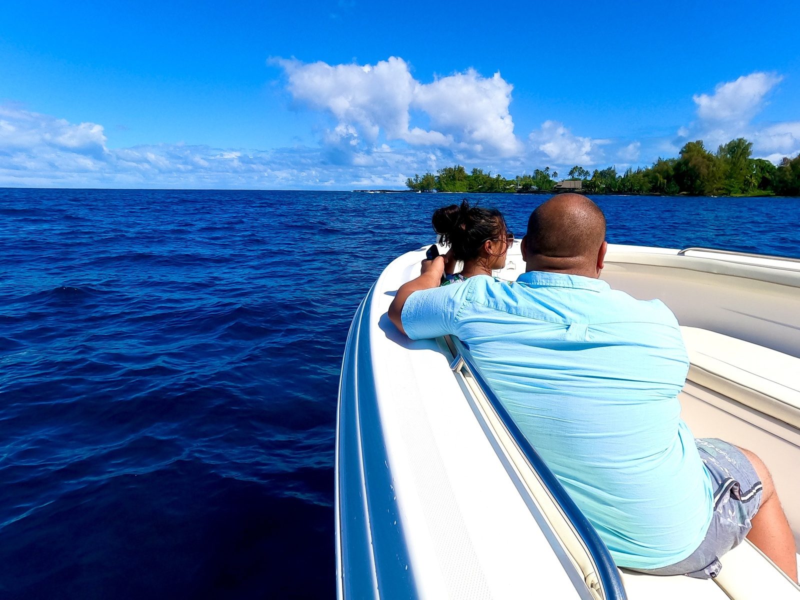Two guests leaning on the bow of a white boat in deep blue water, looking toward a green, palm-lined shoreline