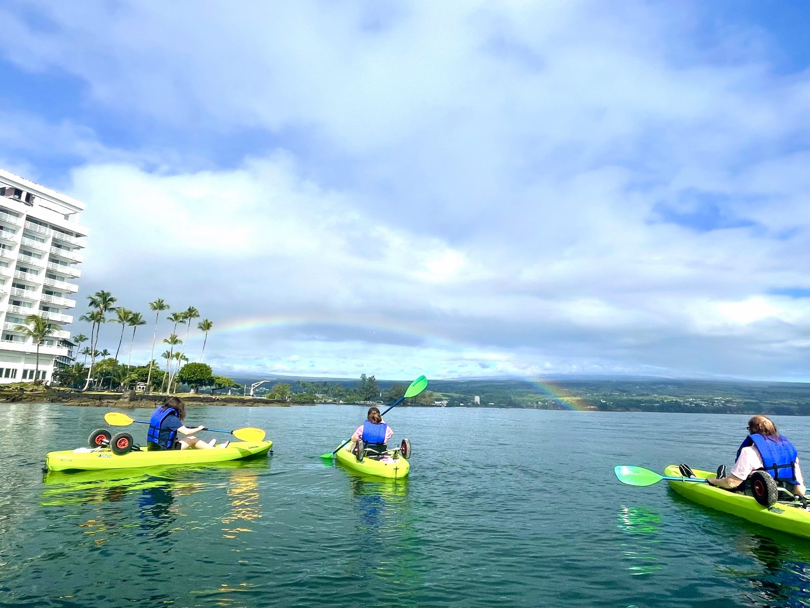 Three kayakers in life jackets paddling Hilo Bay beneath a rainbow, with a tall bayfront hotel and palms on the left