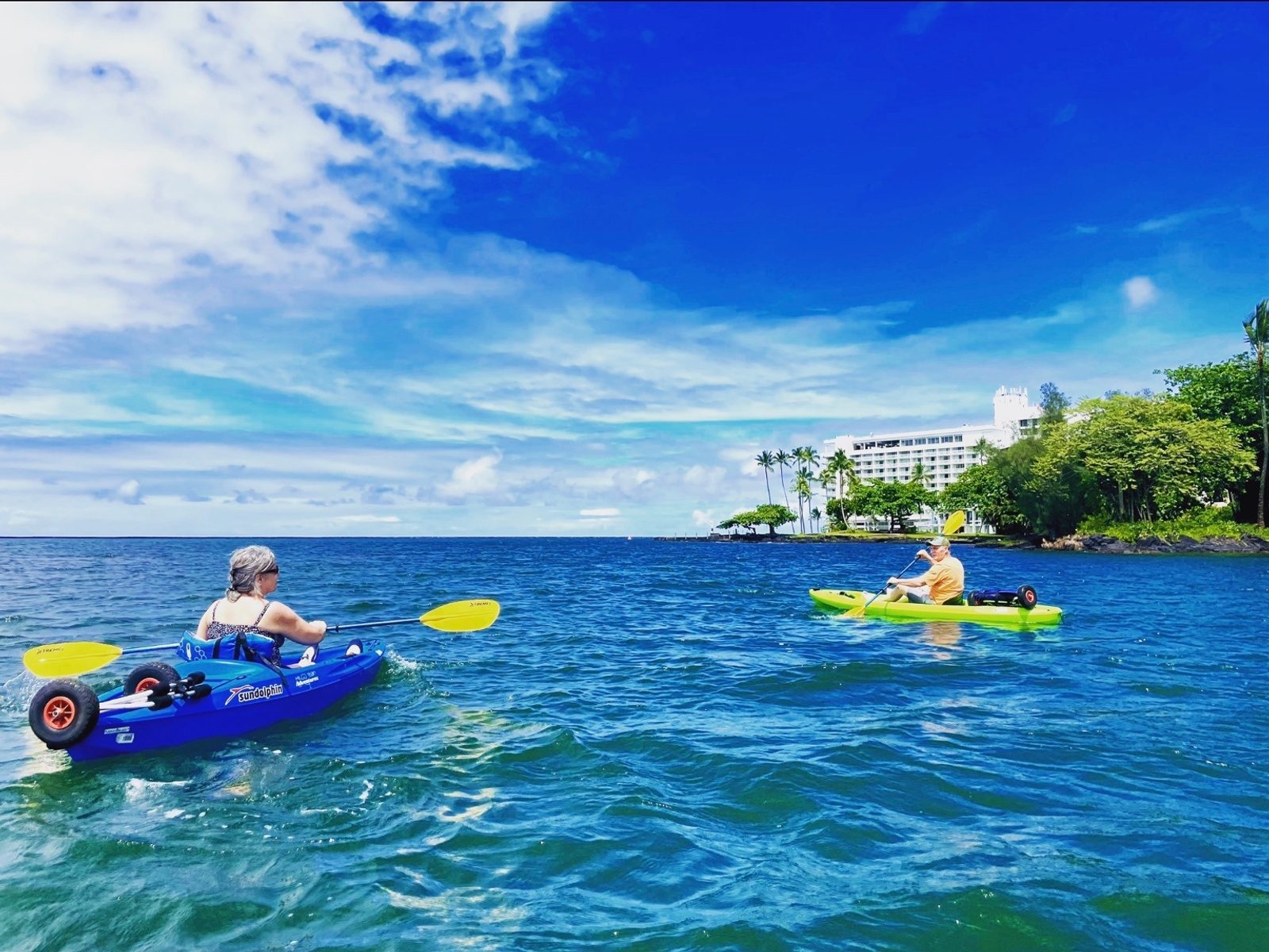 Two kayakers in blue and green kayaks on choppy blue water off a palm-fringed point on Hilo Bay
