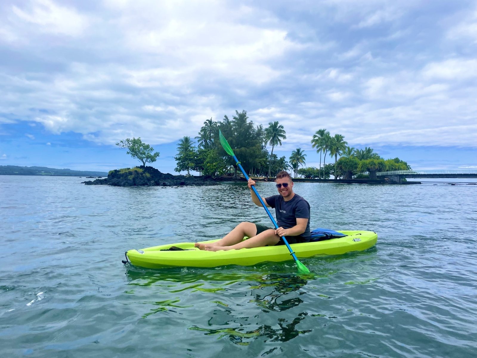 A paddler in a green kayak on calm water in front of Coconut Island, a small palm-covered islet linked to shore by a footbridge