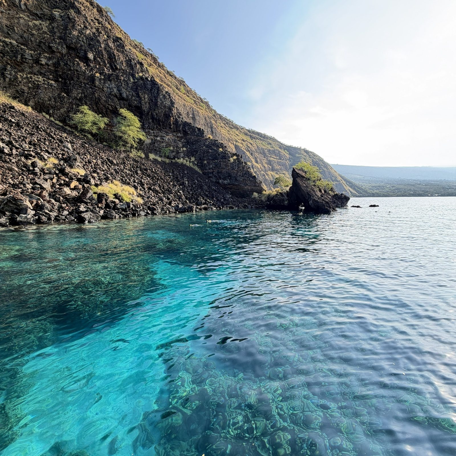 Glassy blue water over a shallow reef beside a steep cliff and a rocky point, with a few snorkelers near the shoreline