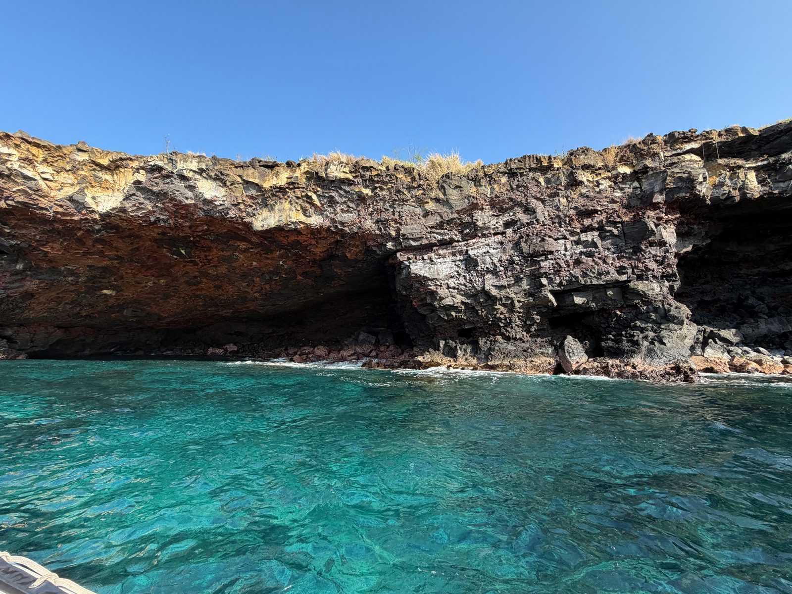A wide, low sea cave cut into a layered lava cliff above clear turquoise water