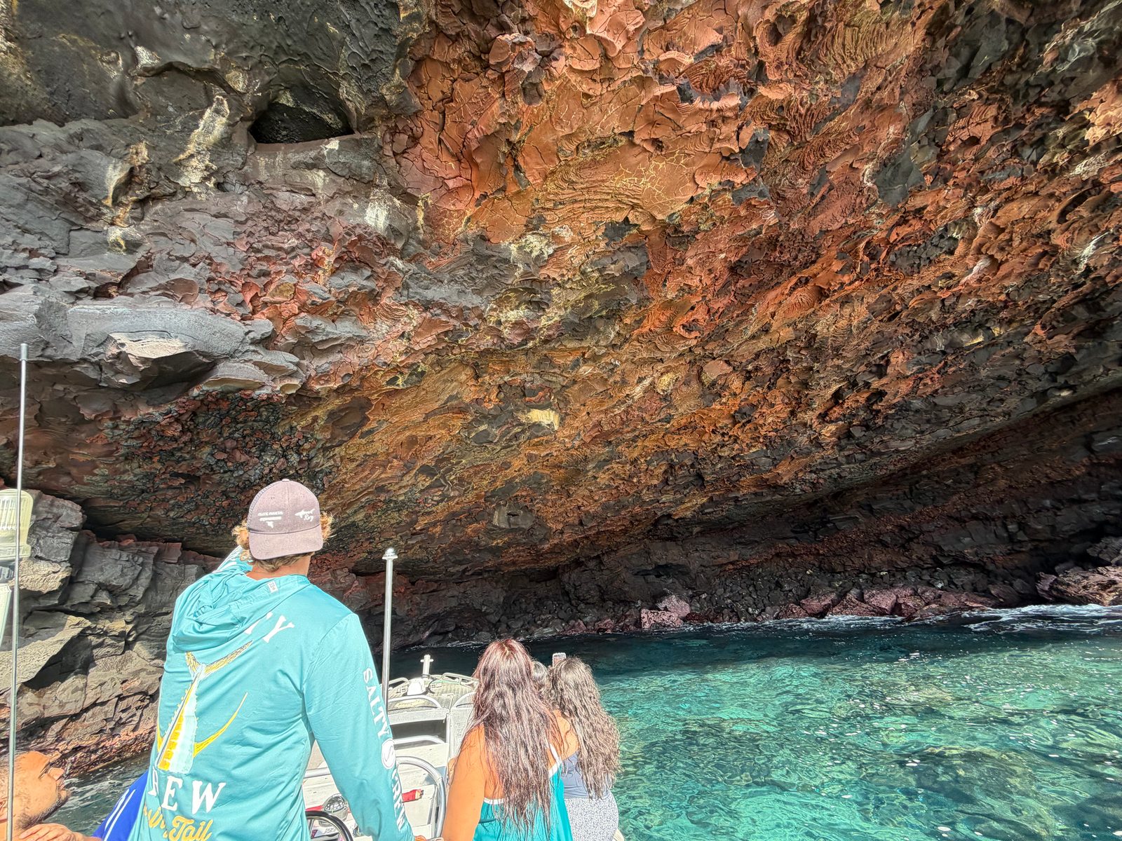 Guests on the bow of the boat looking up at the red, orange and black rock ceiling of a sea cave over clear turquoise water