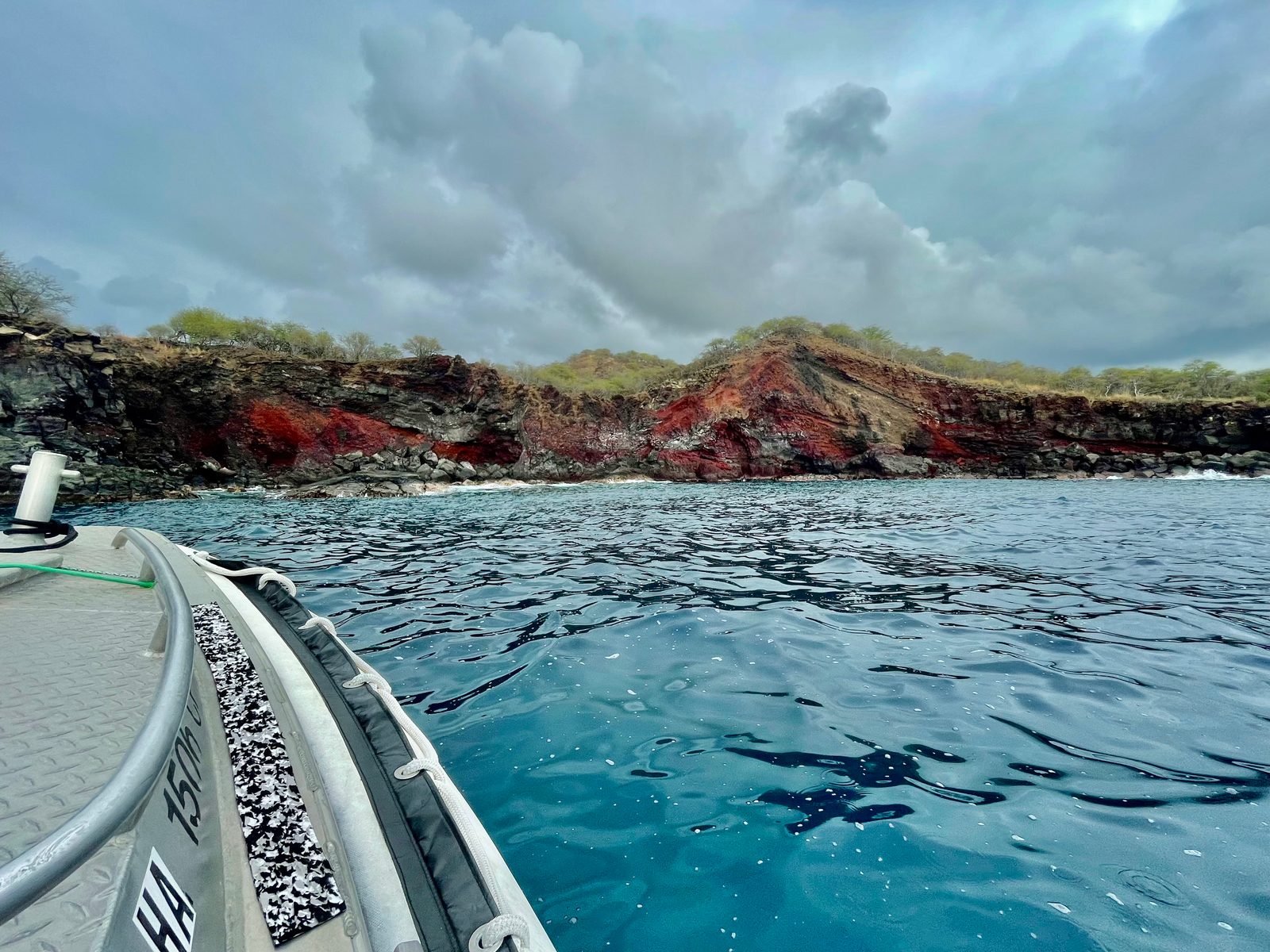 The bow of the inflatable boat pointing at a coastline of red and black volcanic cliffs under heavy gray clouds