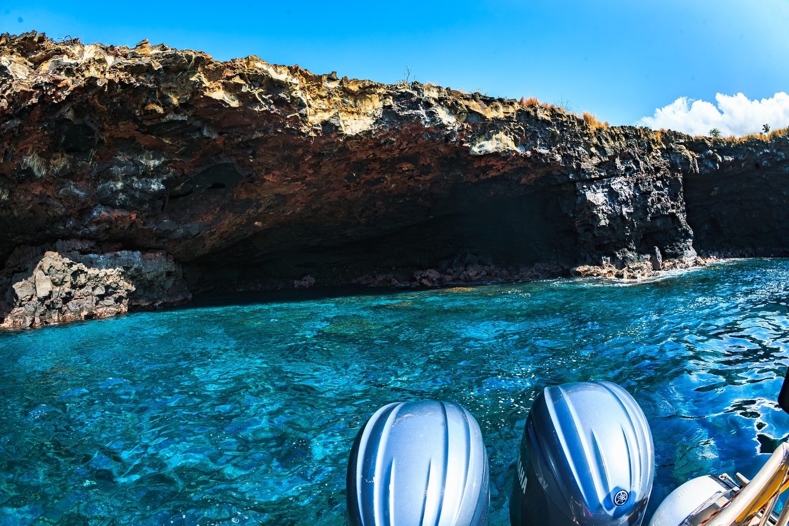 Twin outboard motors in the foreground as the boat idles beside a sea cave in a jagged lava sea cliff