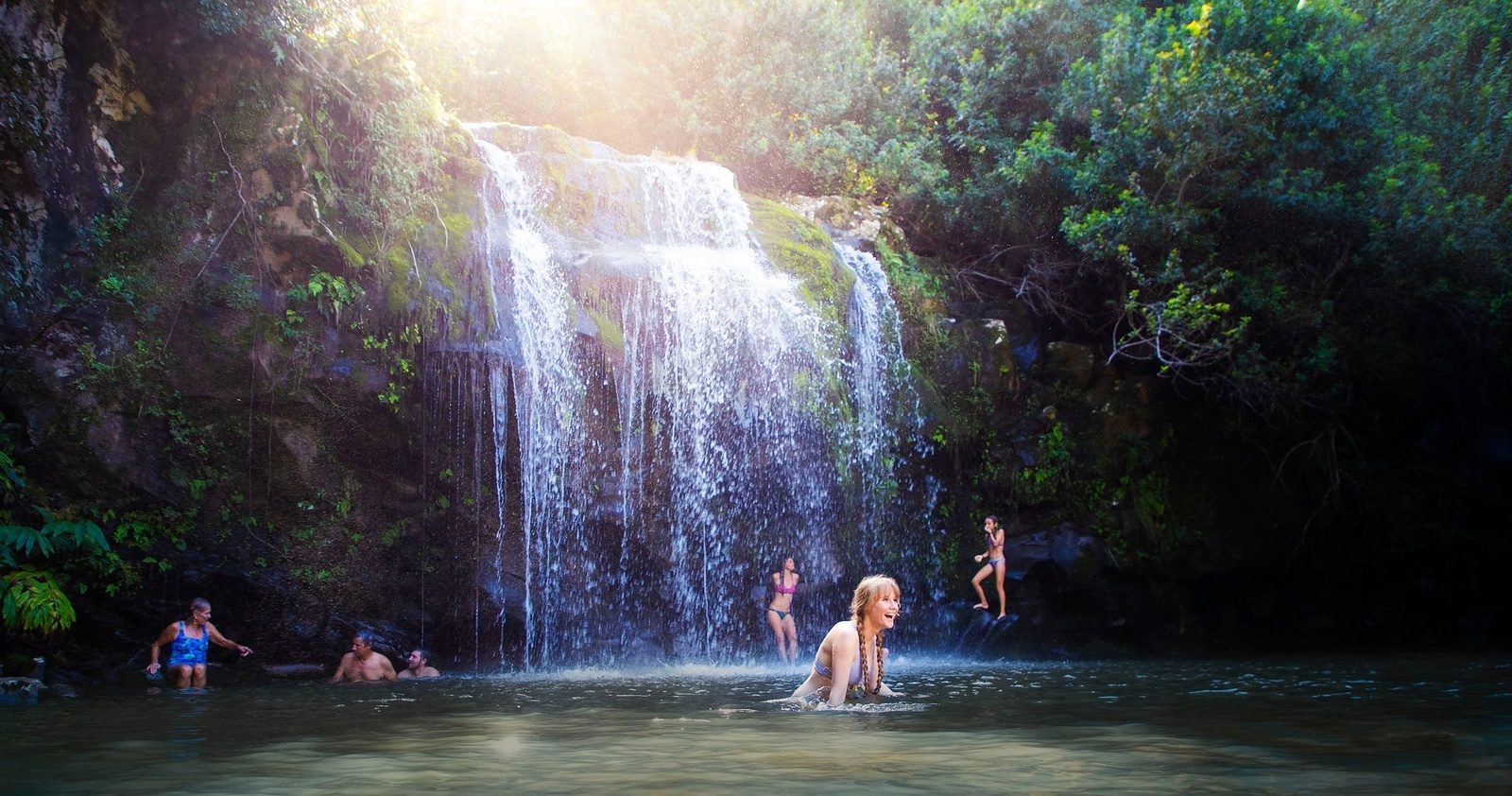 Swimmers laughing in a pool below a wide waterfall in a sunlit forest, the swim stop on the Zip and Dip day