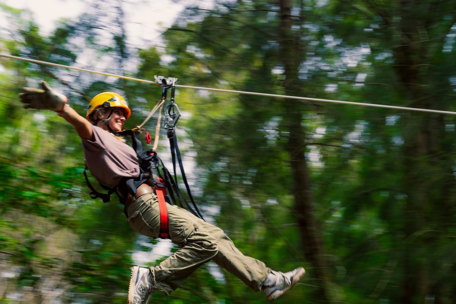 A woman in a yellow helmet flying along a zipline with one arm out, the forest a green blur behind her