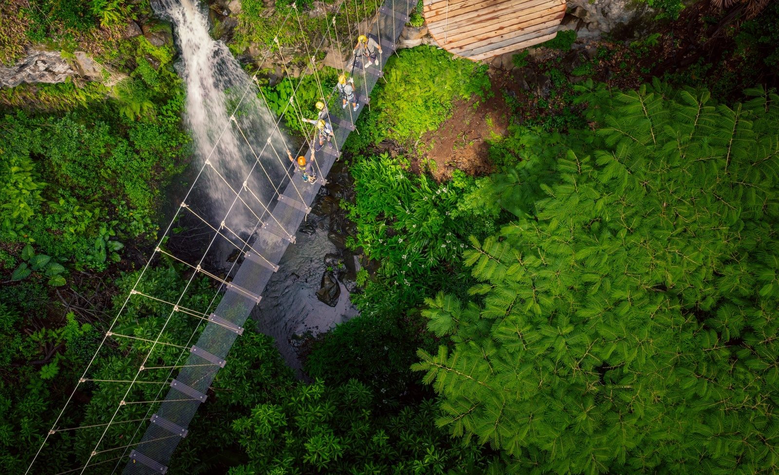 Overhead view of five zipliners in helmets crossing a narrow suspension bridge above a waterfall and a rocky stream in dense green forest