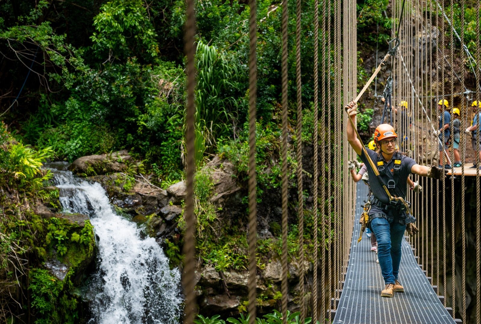 A guide in an orange helmet walking a cable suspension bridge beside a white-water waterfall, with guests waiting on a platform behind