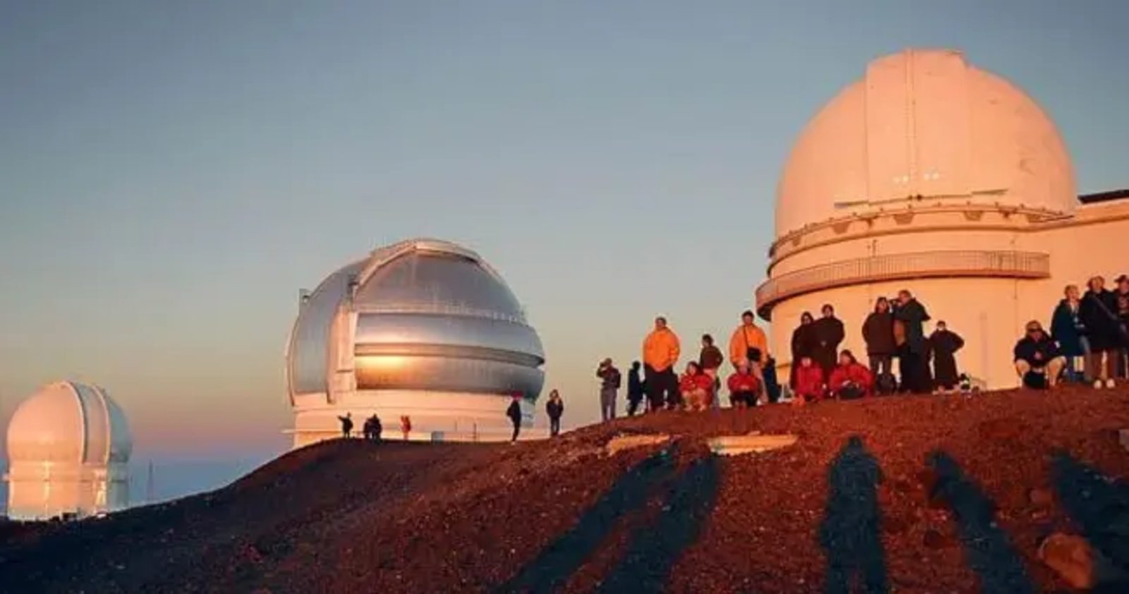 A crowd of people in warm jackets standing on reddish cinder beside two large observatory domes in golden sunset light