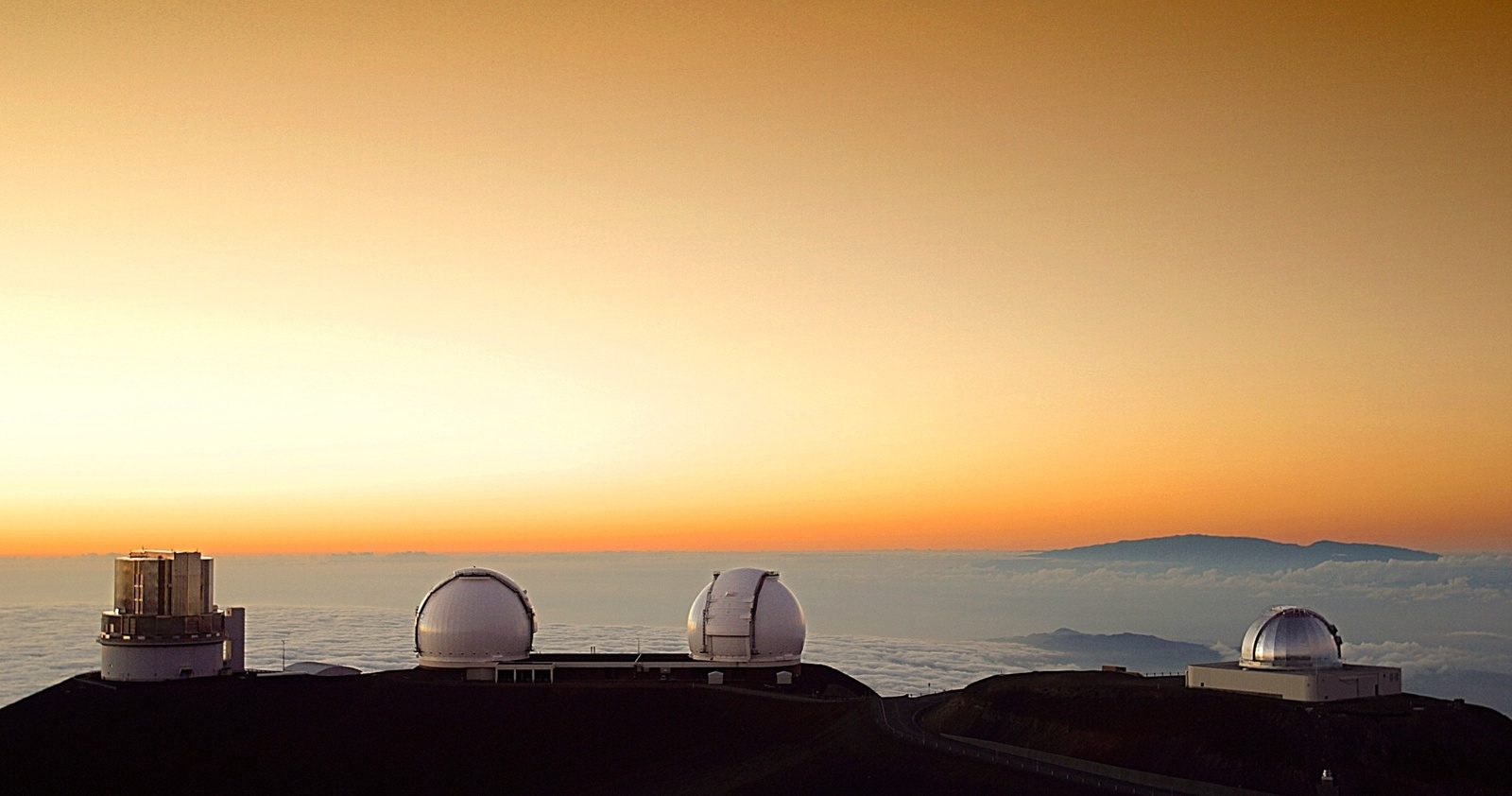 A row of white and silver telescope domes on a dark ridge above a sea of clouds, with another mountain on the horizon under an orange sky