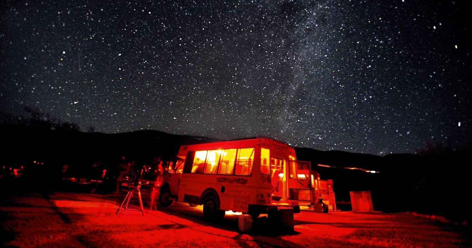 A tour van glowing with red interior light under a starry sky, with a telescope on a tripod set up beside it