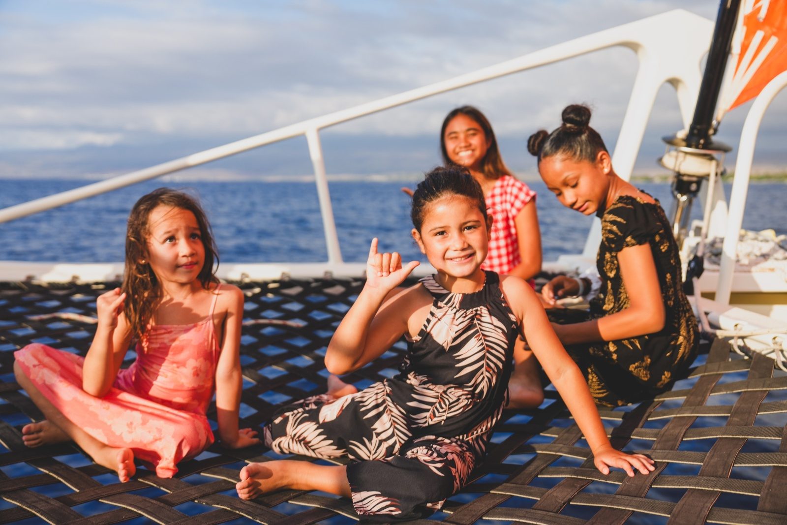 Four girls in island dresses sitting on the catamaran's trampoline net in the evening sun, one throwing a shaka