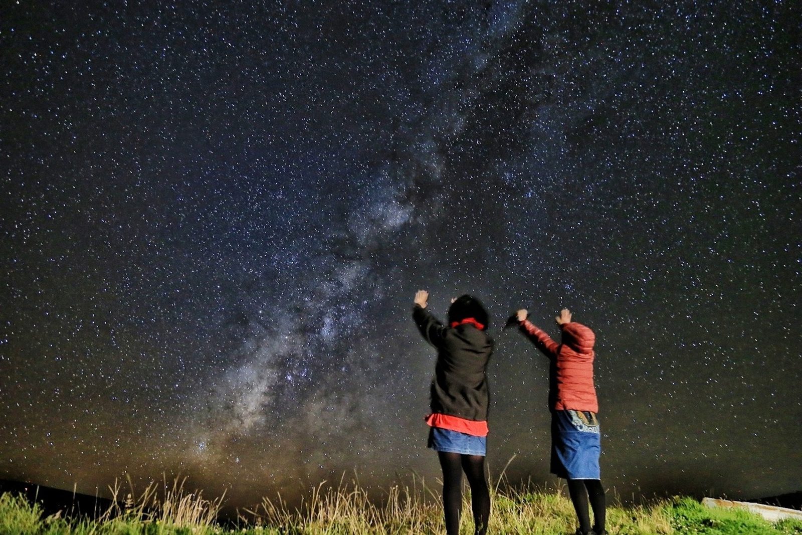 Two people in warm jackets raising their arms toward the Milky Way in a sky full of stars above a grassy rise