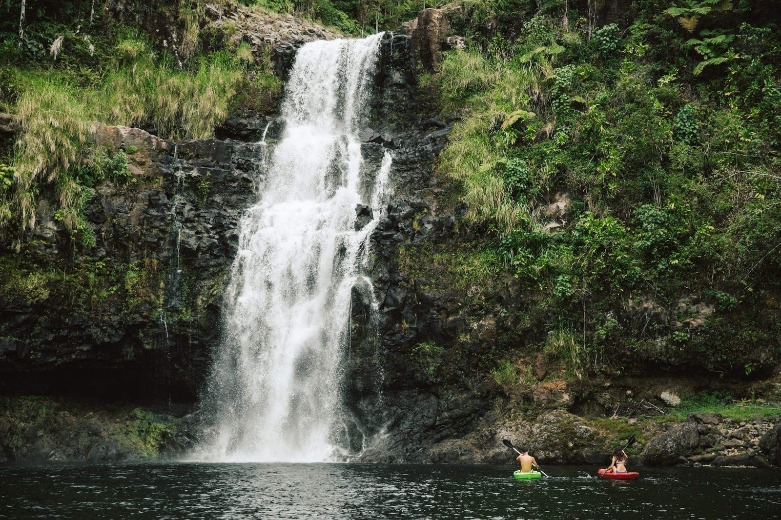 Two people paddling small inflatable boats in a dark pool at the base of a tall waterfall pouring over black lava rock